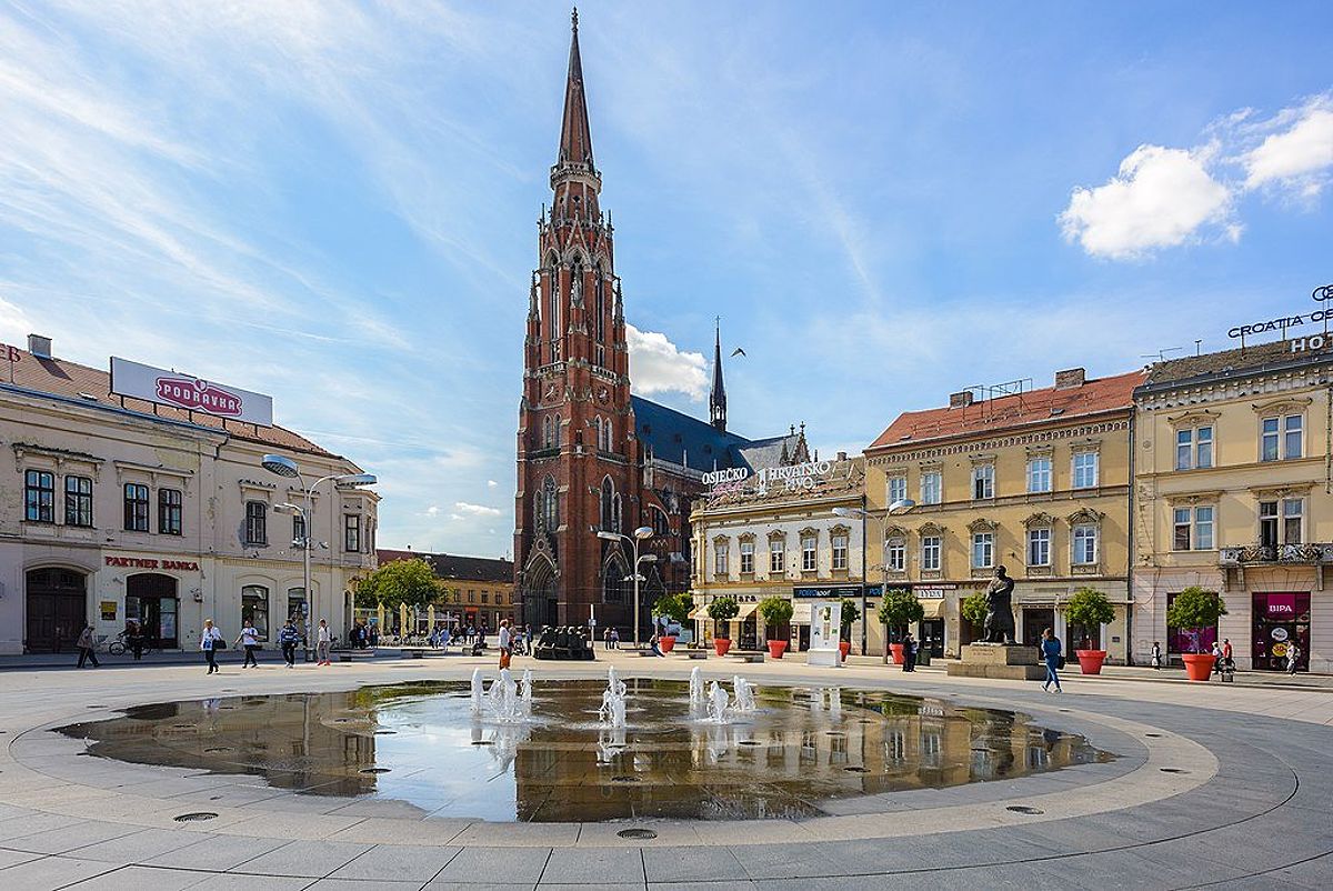 The main square with water jets rising slightly out of a ground fountain and a red church steeple prominent in Osijek, in the Baranja Region.