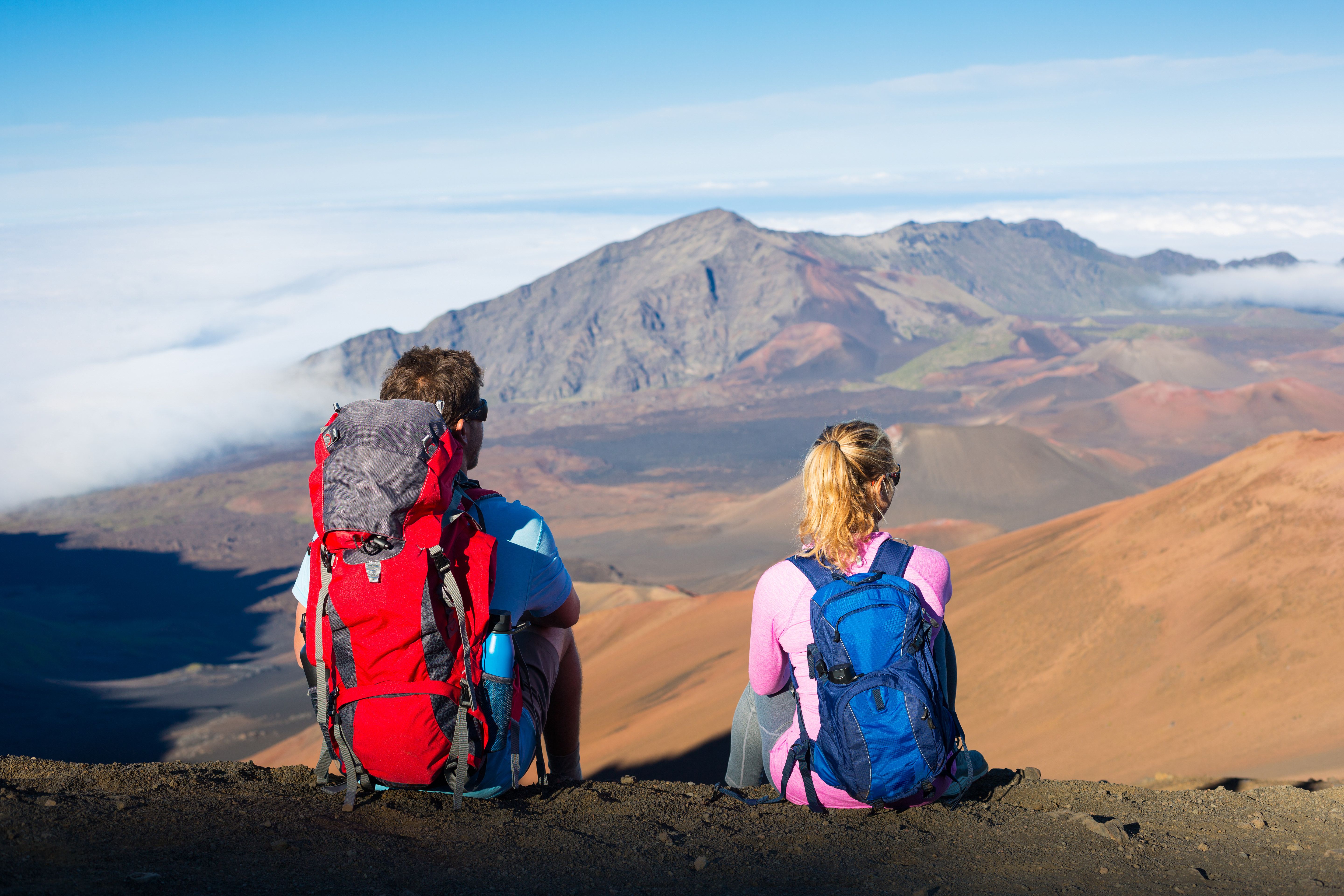 Two hikers sit on the edge of a crater looking out at a volcanic landscape